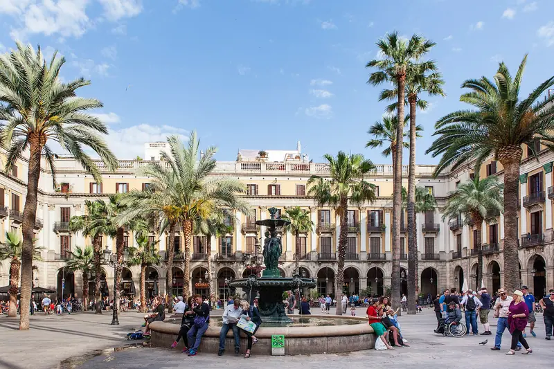 Plaza Real de Barcelona con palmeras, una fuente central y soportales de fachadas amarillas, con gente sentada y paseando