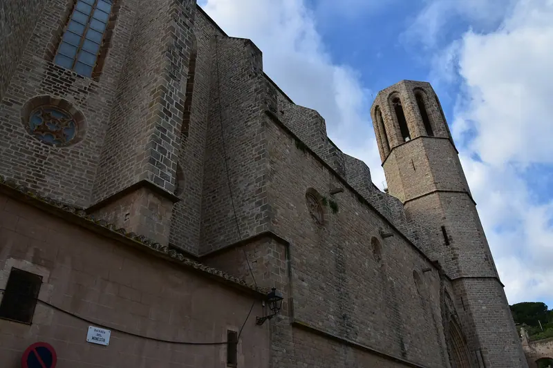 Muro de piedra y torre campanario del monasterio de Pedralbes vistos desde abajo, con cielo azul y nubes
