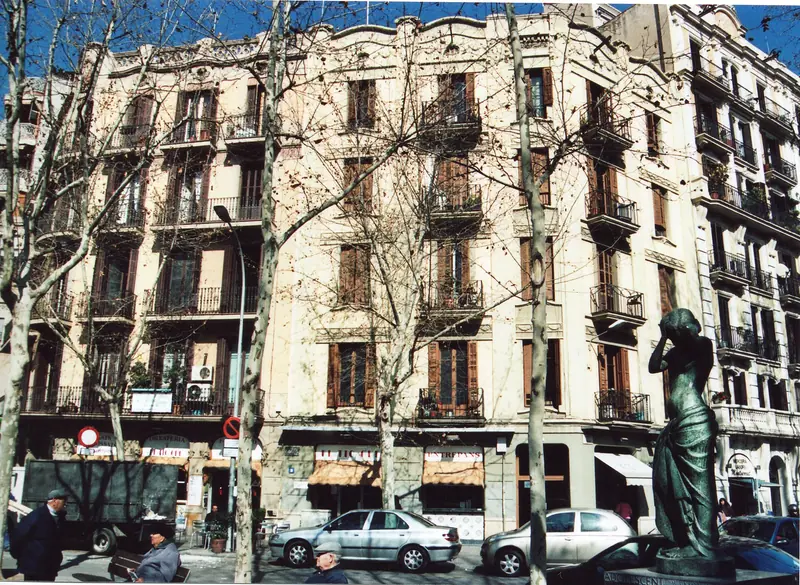 Edificios antiguos con balcones en la Rambla del Poblenou, entre plátanos sin hojas, con coches aparcados, algunas personas sentadas y una estatua a la derecha