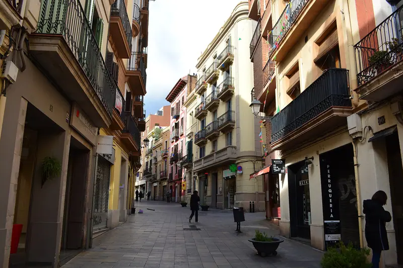 Calle peatonal del centro de Sarrià con balcones de hierro, comercios y fachadas de colores, y algunos viandantes