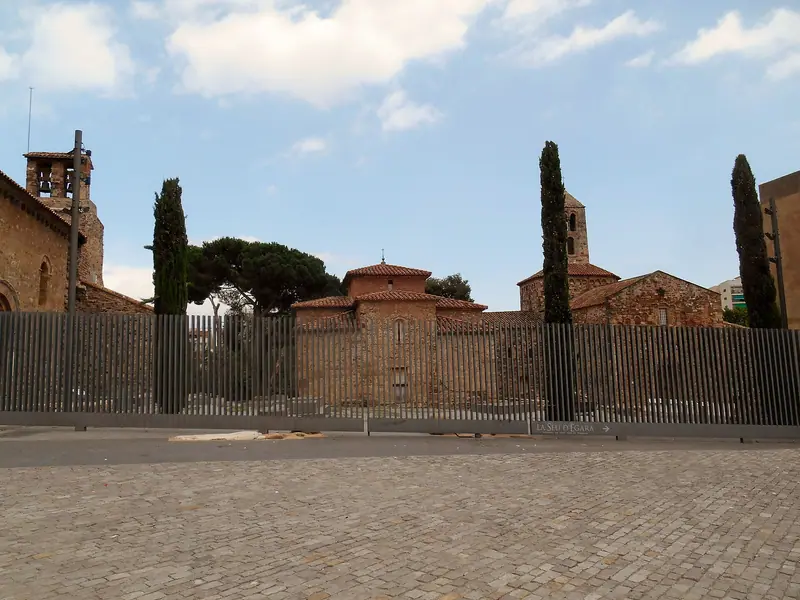 Conjunto de iglesias románicas de la Seu d'Ègara tras una valla metálica, con cipreses y una plaza empedrada en primer plano